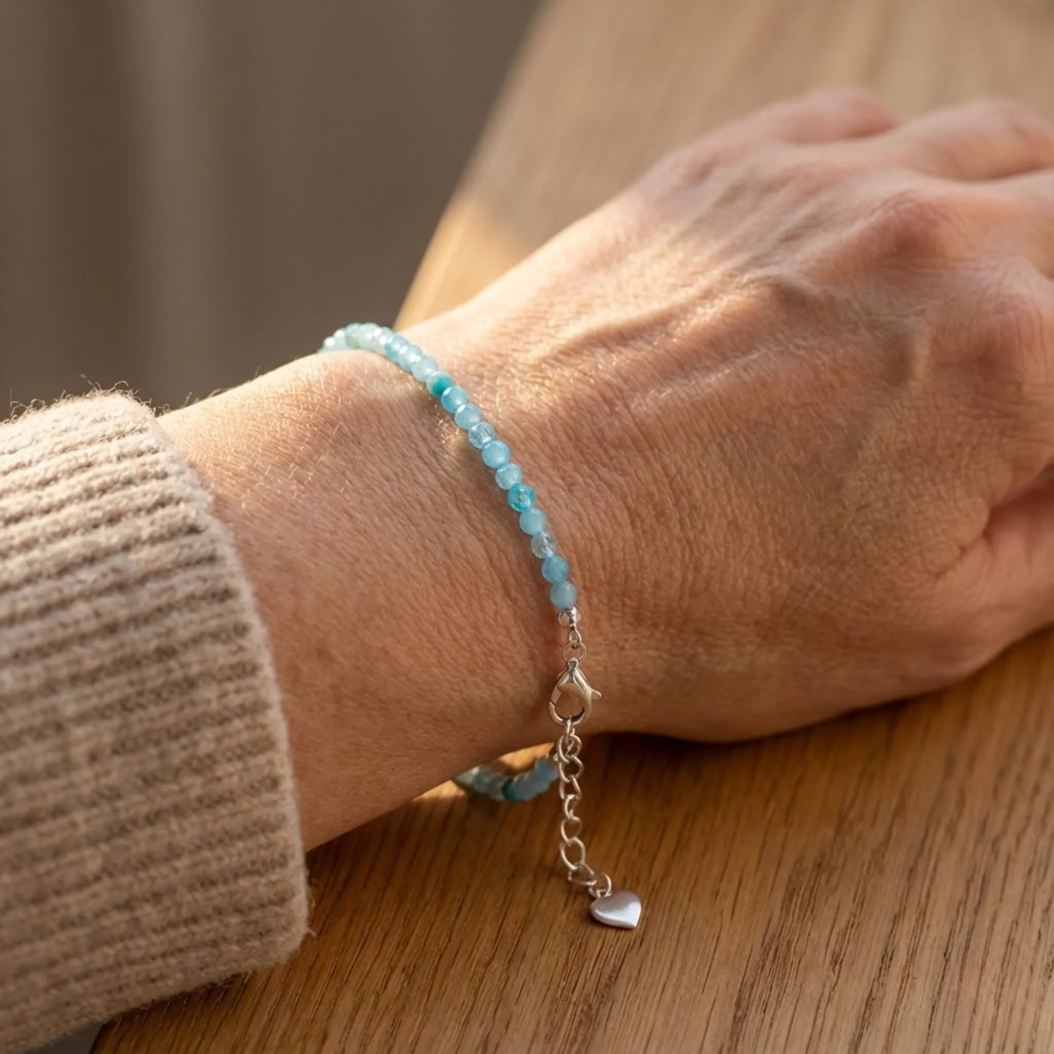Close-up of a hand wearing a Essential silver bracelet crafted with amazonite gemstone beads in calming green-blue shades bracelet with a heart charm on a wooden surface