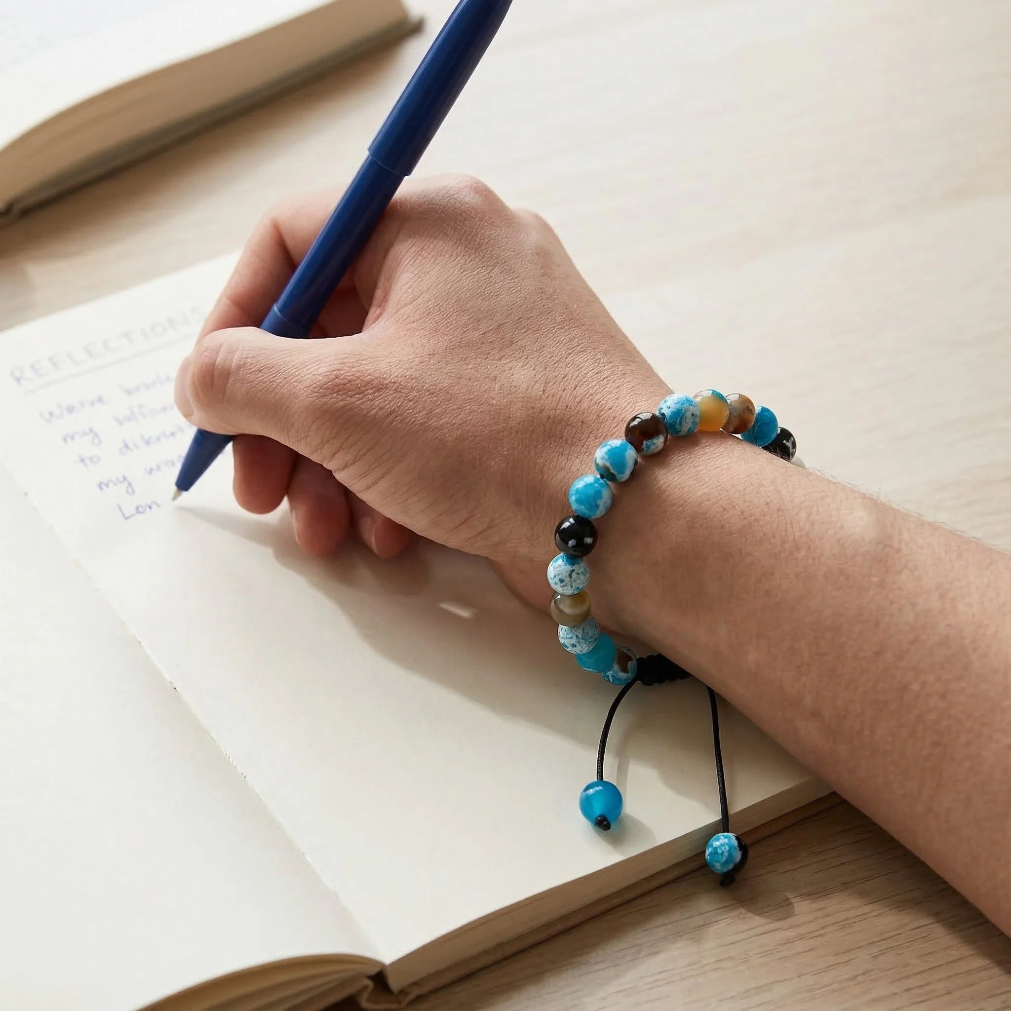 Hand writing in a notebook with a blue pen, wearing a colorful beaded bracelet.