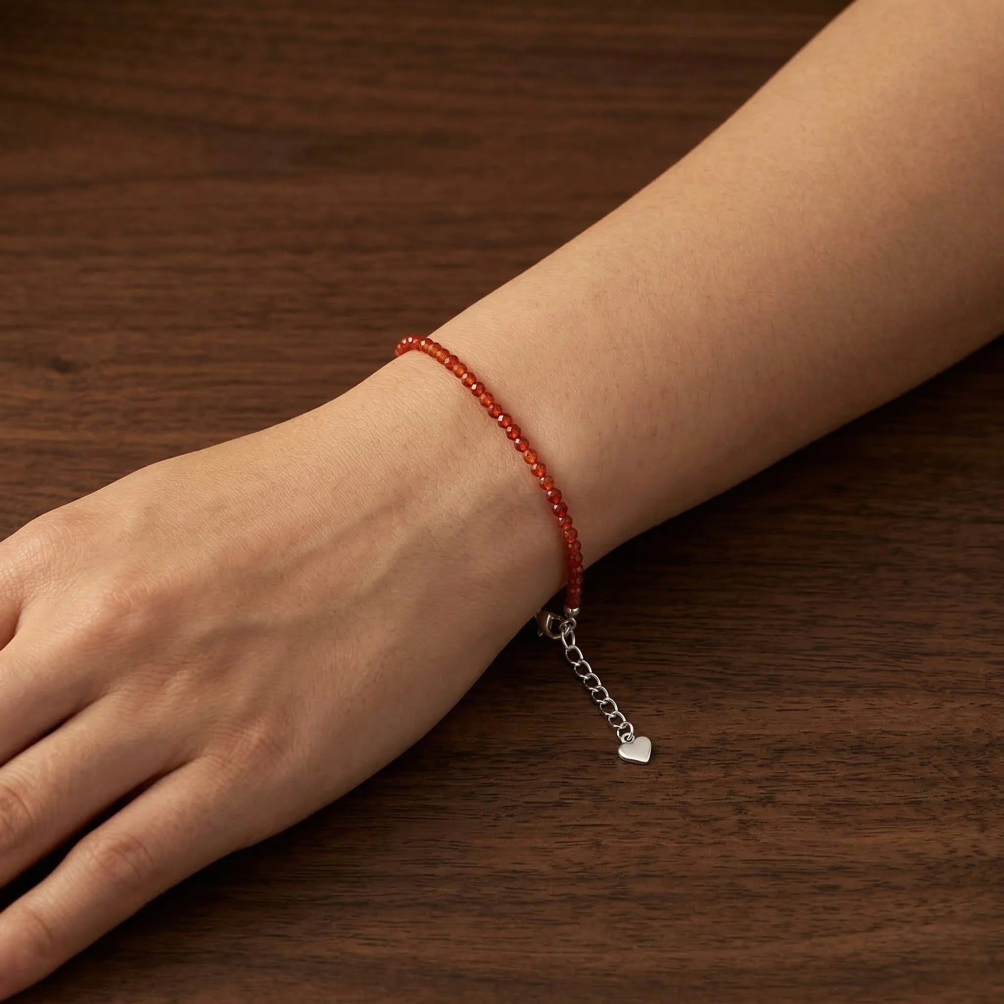Hand wearing a red beaded bracelet with a silver charm on a wooden surface