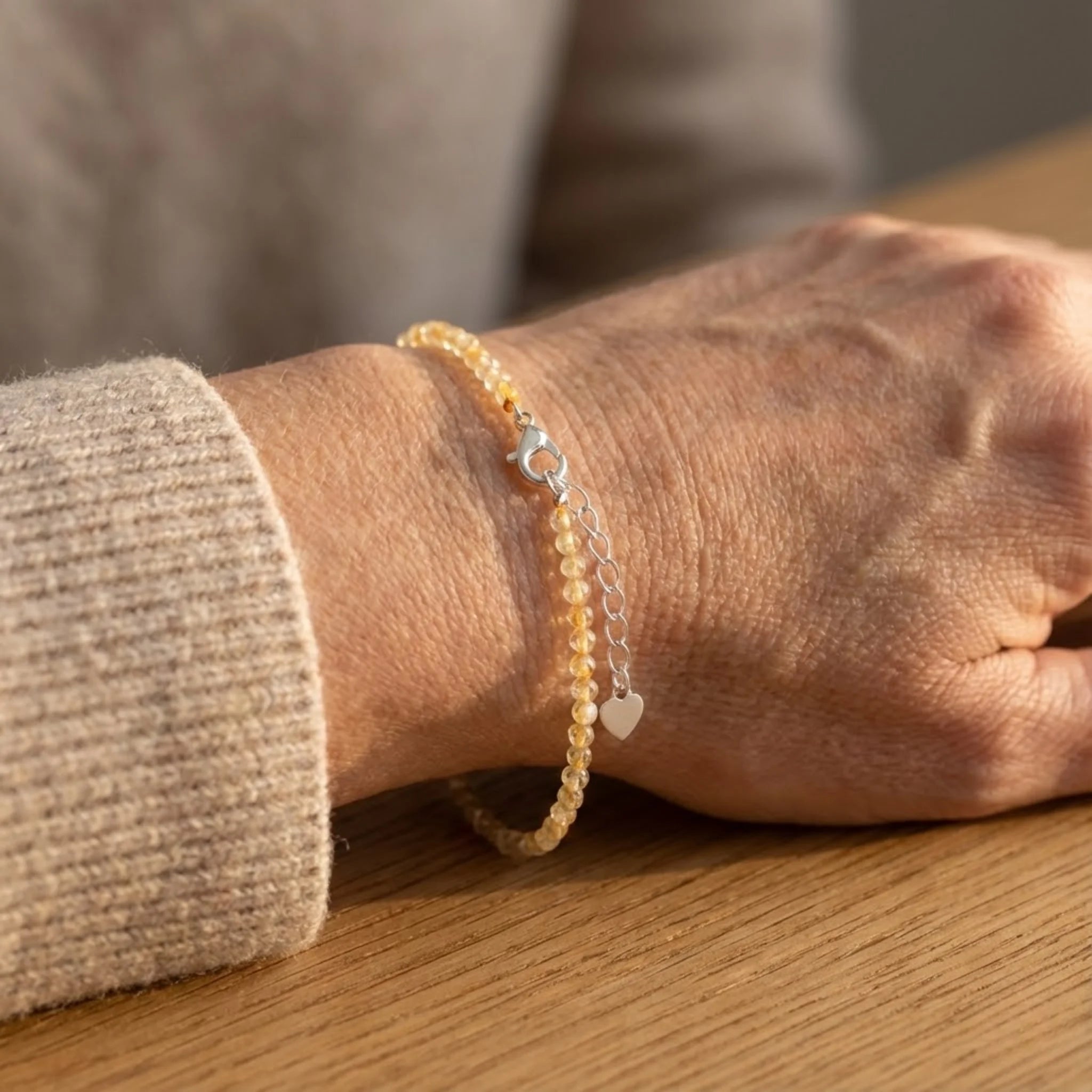 Close-up of a wrist wearing a yellow beaded Essential silver bracelet featuring golden rutilated quartz with natural golden inclusions bracelet with silver charms on a wooden surface.
