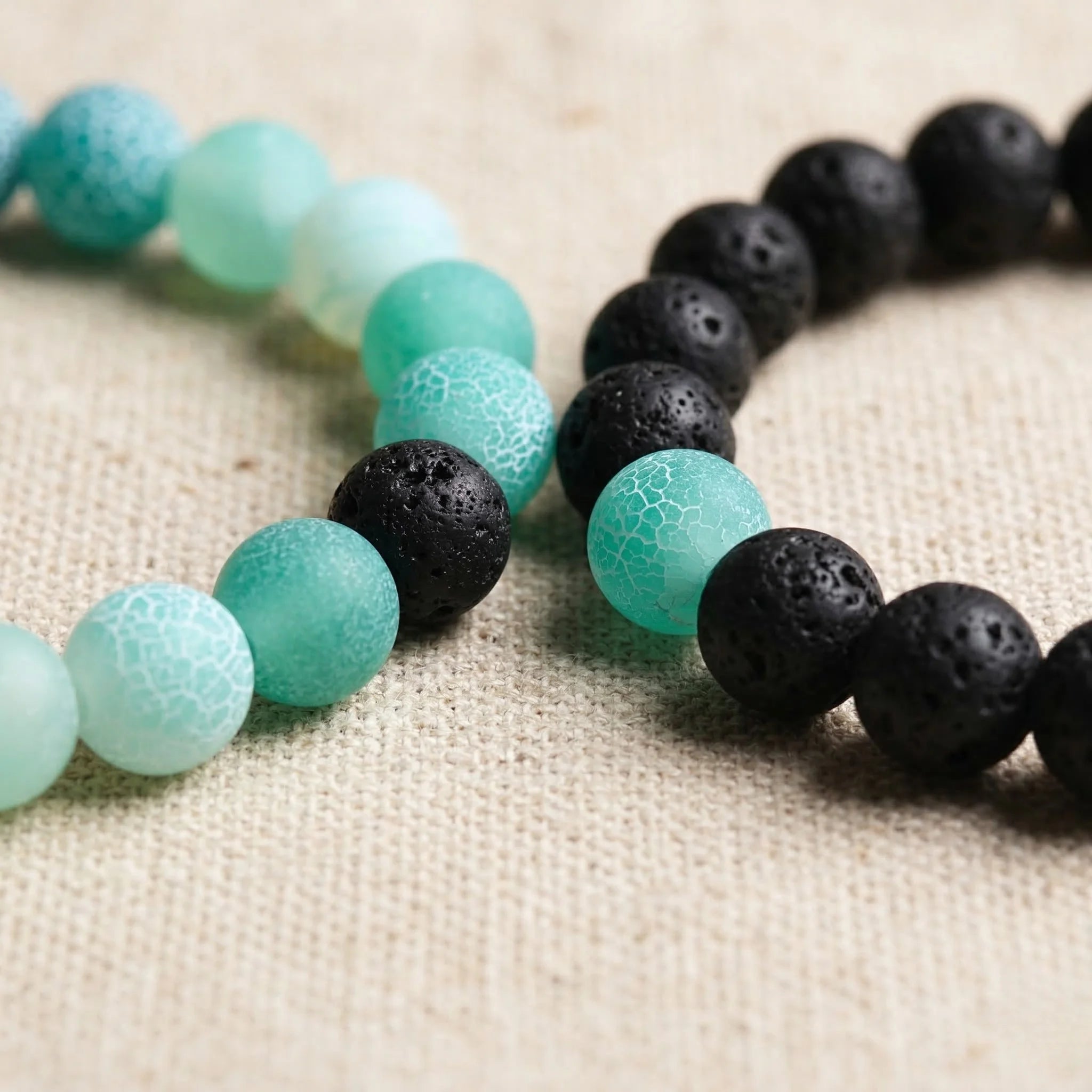 Two beaded bracelets, one with turquoise beads and the other with black beads, on a textured beige surface.