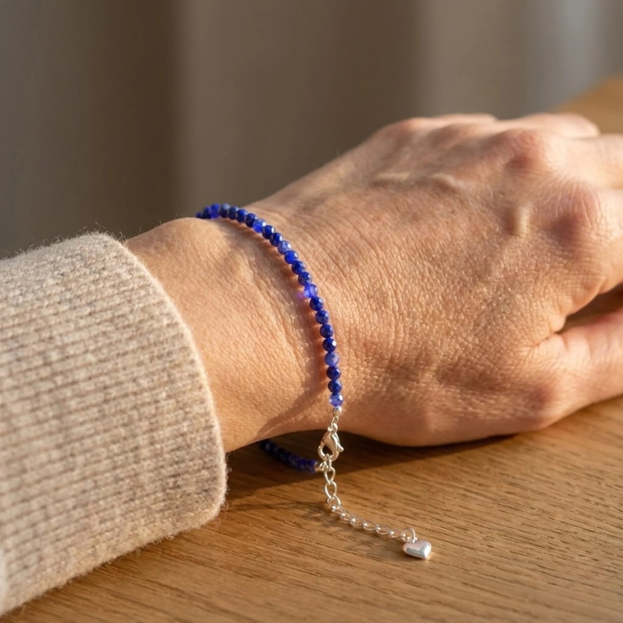 Close-up of a wrist wearing a blue beaded Essential silver bracelet featuring lapis lazuli gemstone beads with deep blue tones bracelet with a silver chain on a wooden surface.