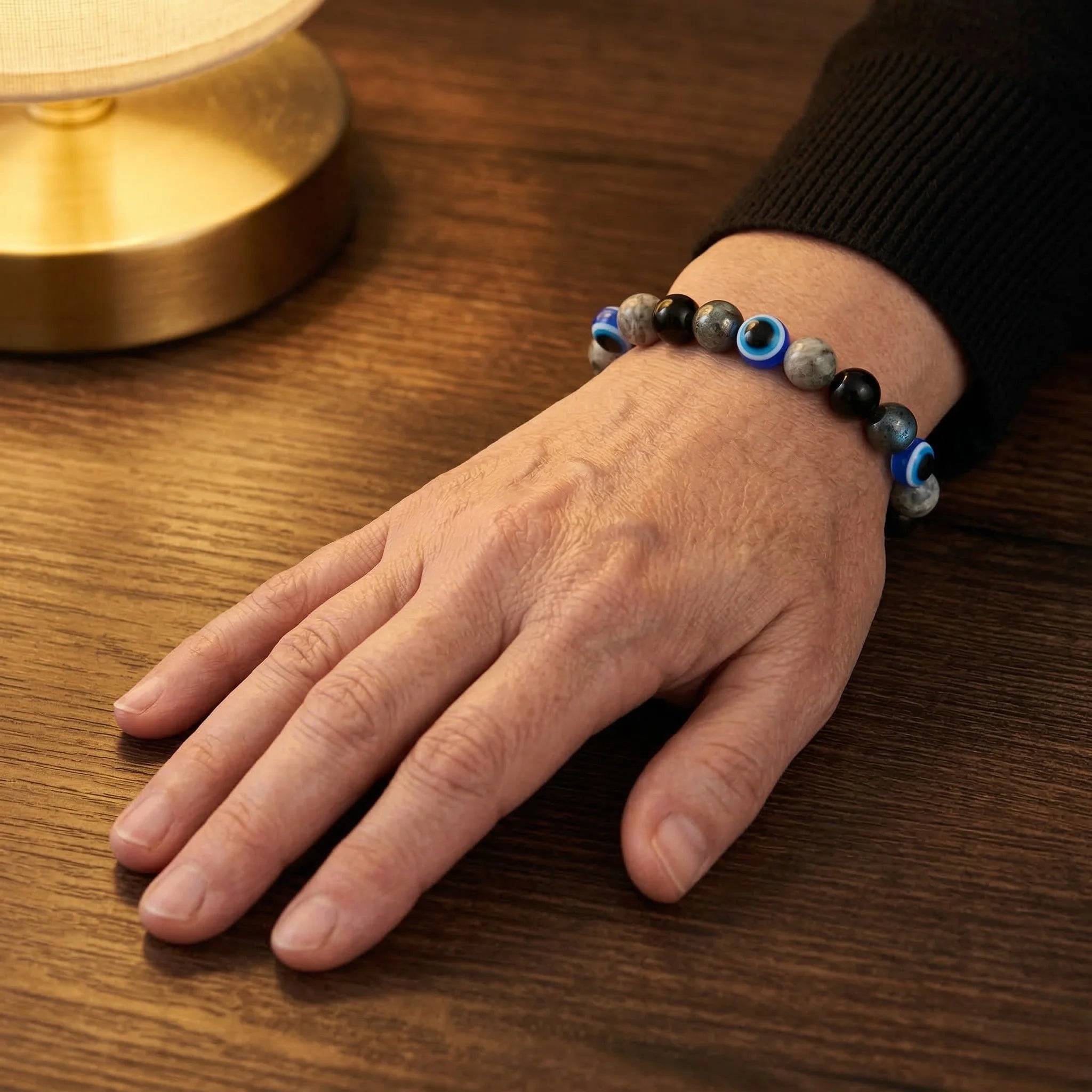 Hand wearing a beaded bracelet on a wooden surface with a lamp in the background