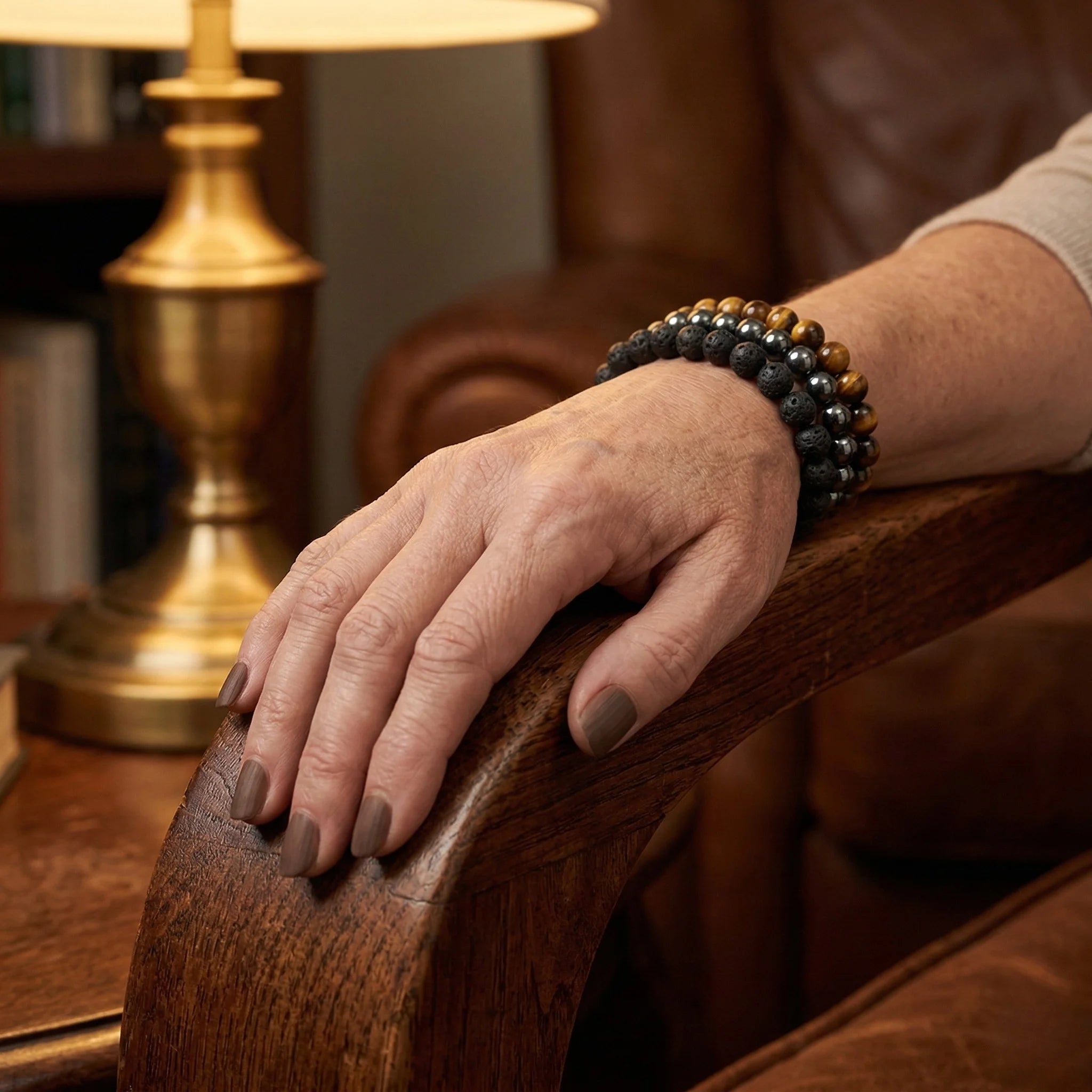 Hand with a bracelet resting on a wooden armrest, with a gold lamp in the background.