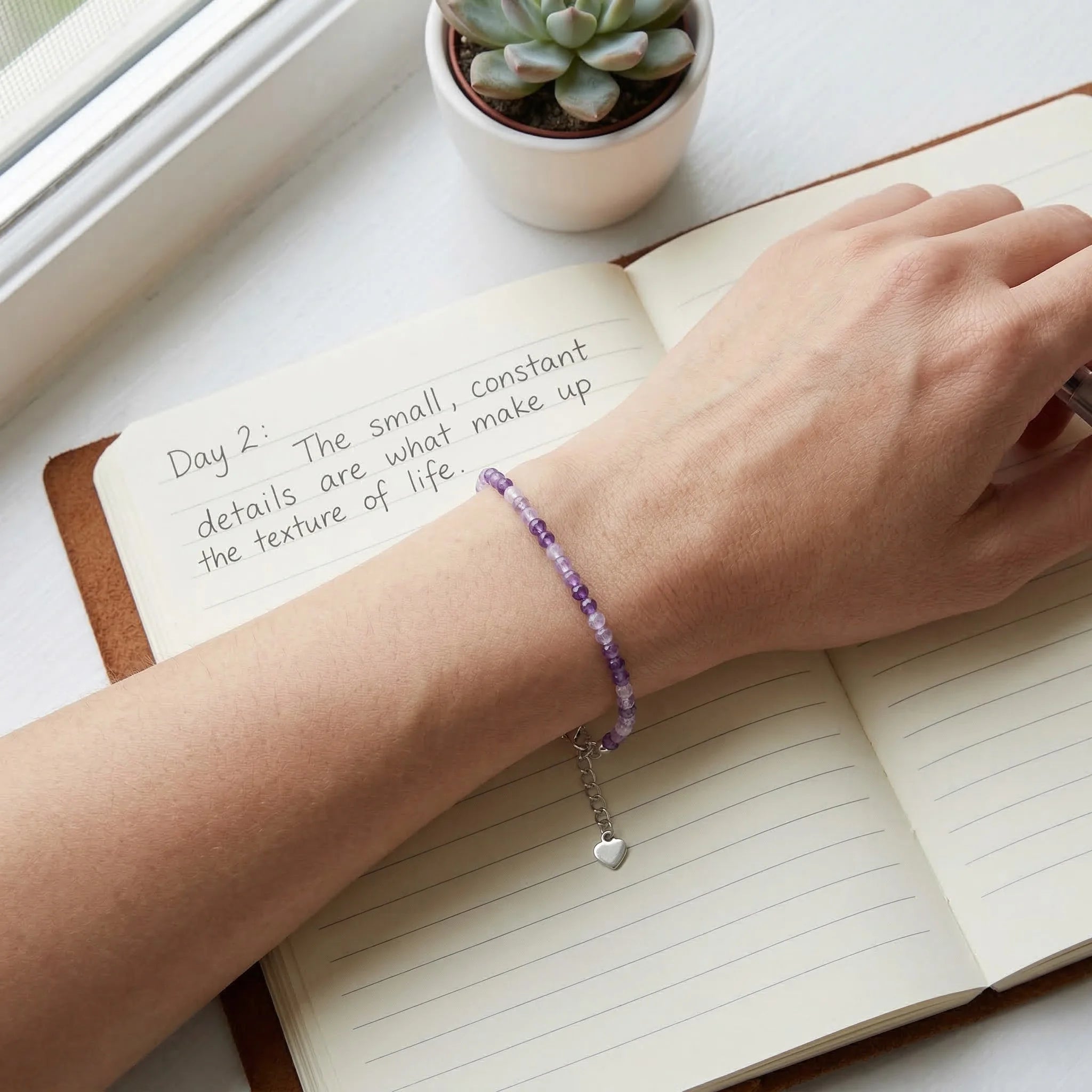 Hand with a purple beaded bracelet on an open notebook with text, next to a small potted plant.