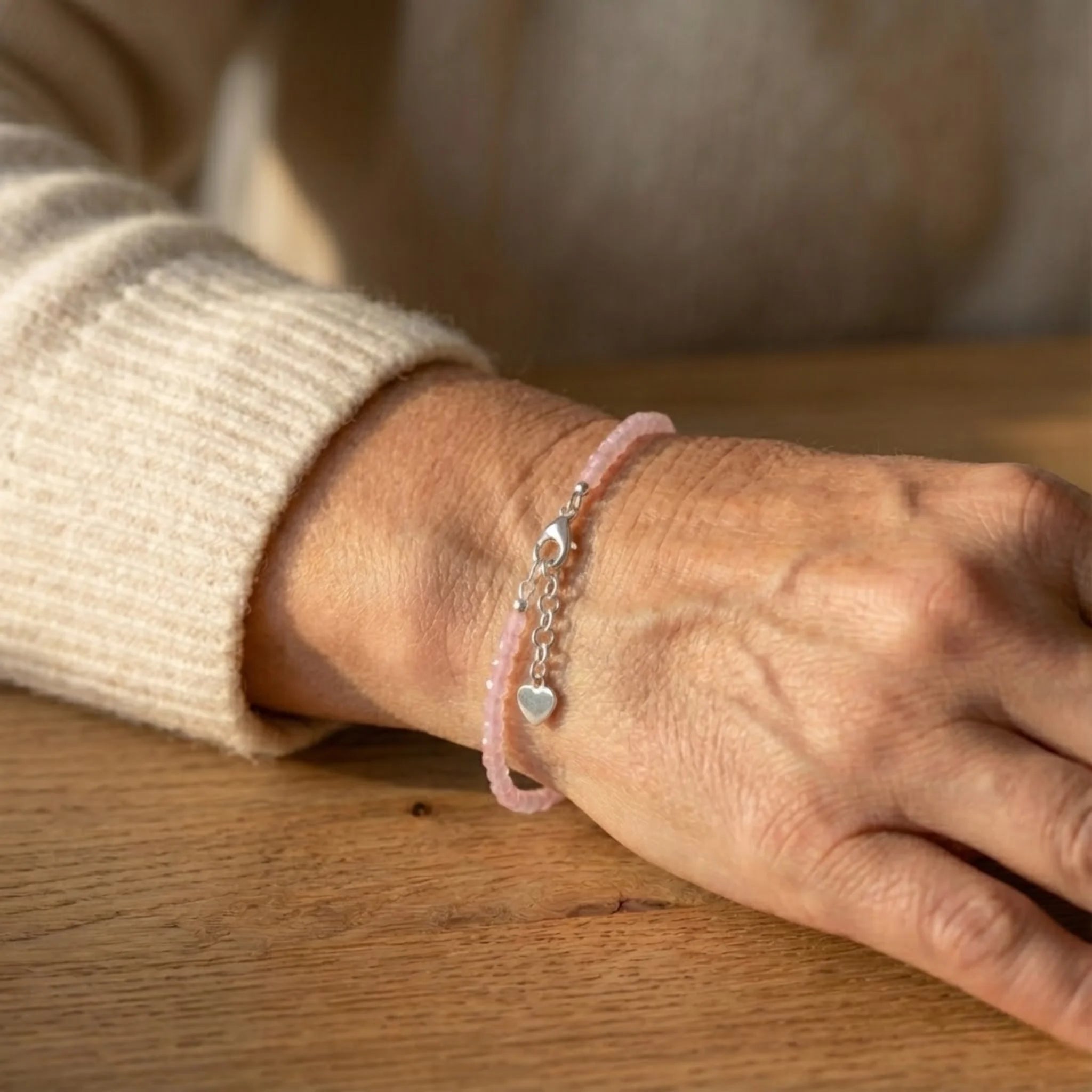 Close-up of a wrist wearing a pink Essential silver bracelet featuring rose quartz gemstone beads for a soft, elegant everyday look bracelet with heart charm on a wooden surface.