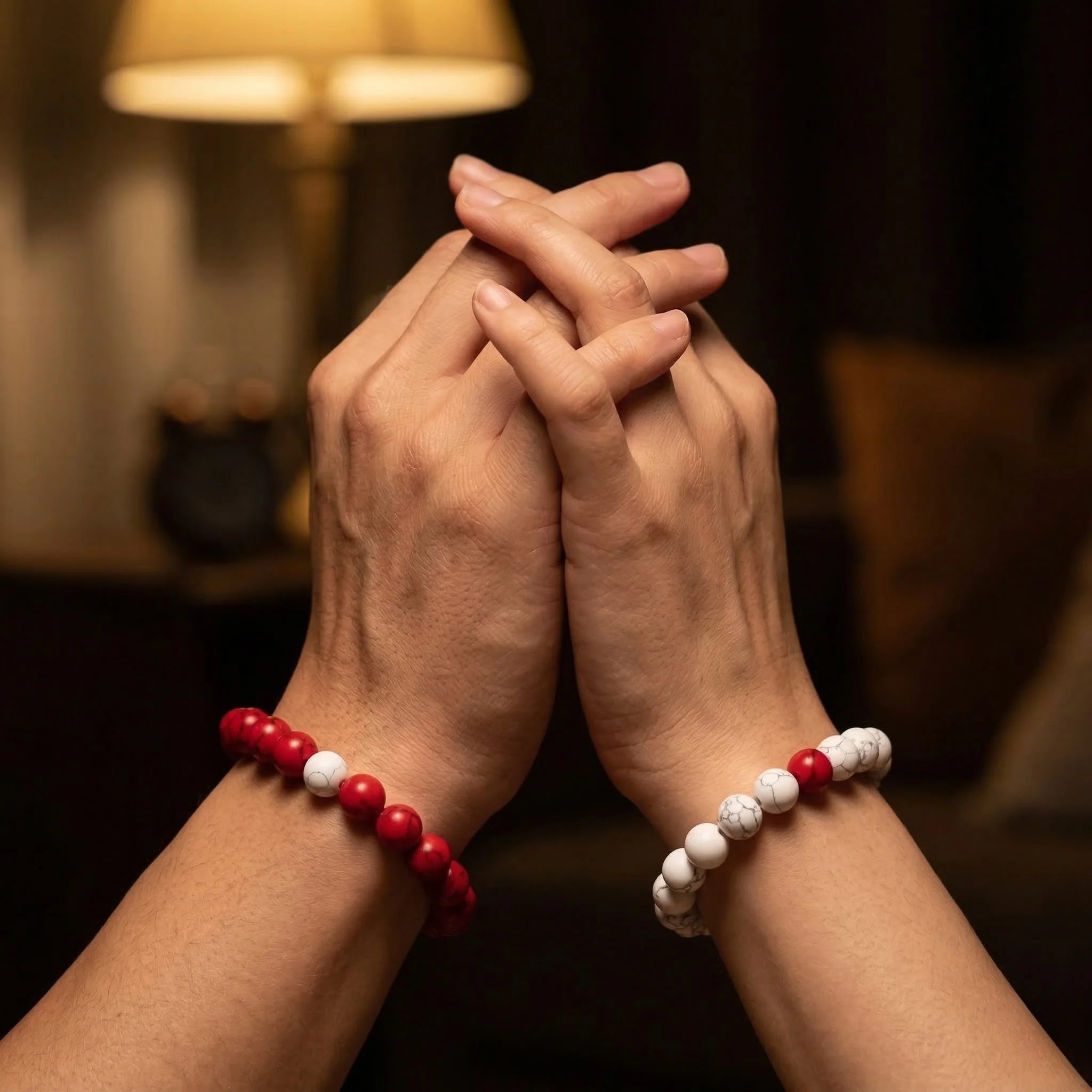 Two hands clasped together wearing red and white beaded bracelets in a dimly lit room.