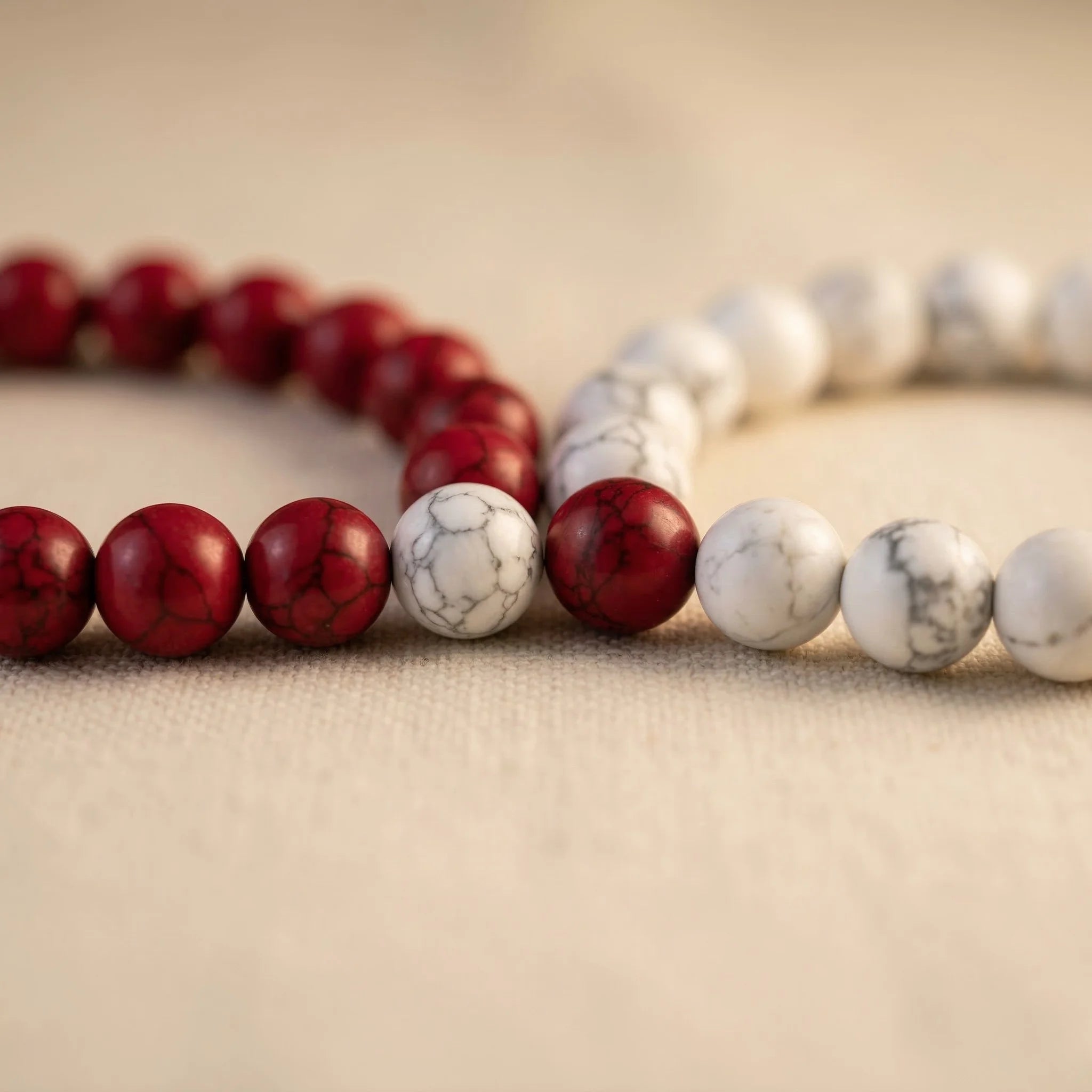 Two beaded bracelets, one red and one white and gray, on a beige fabric background.