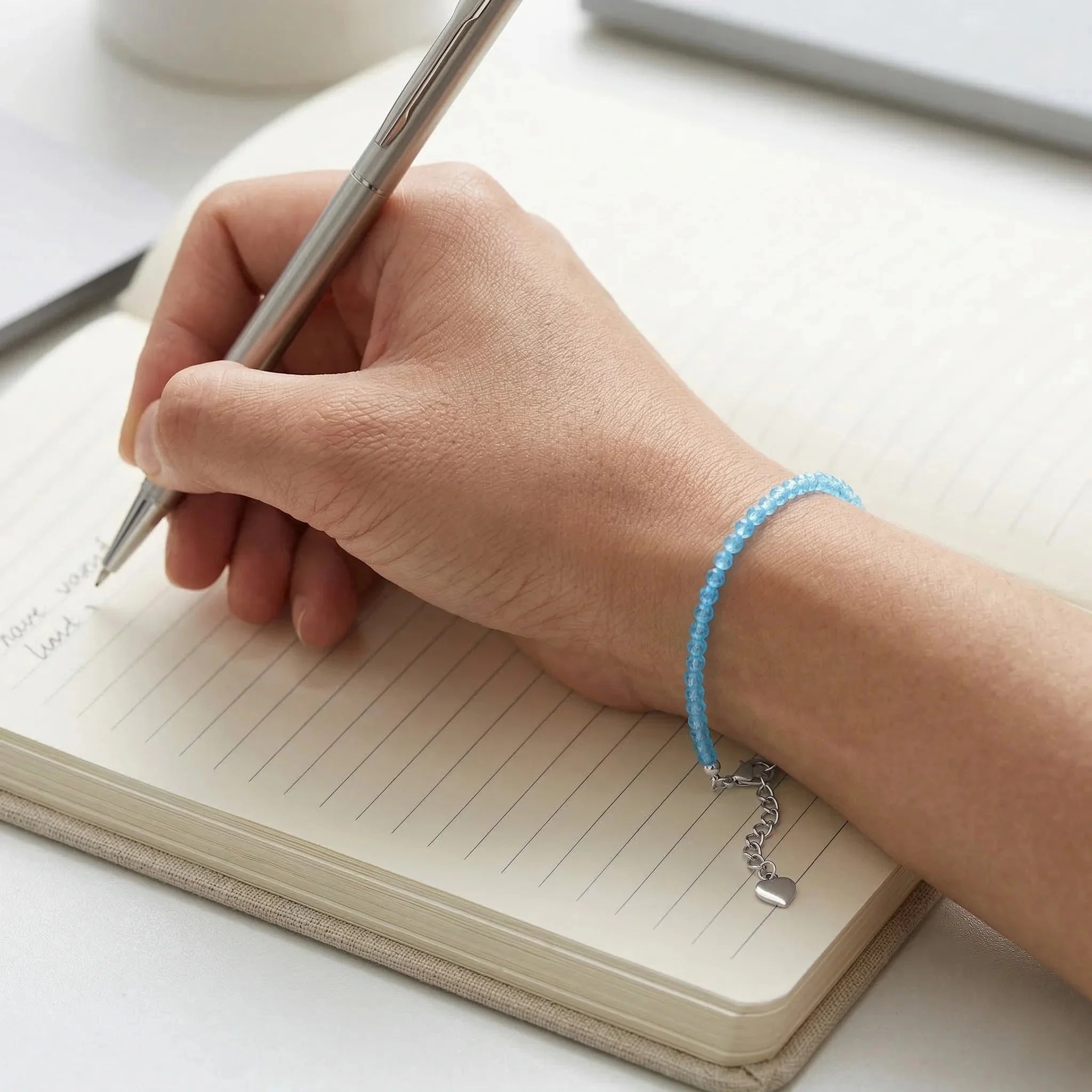 Hand writing in a notebook with a pen, wearing a blue beaded bracelet.