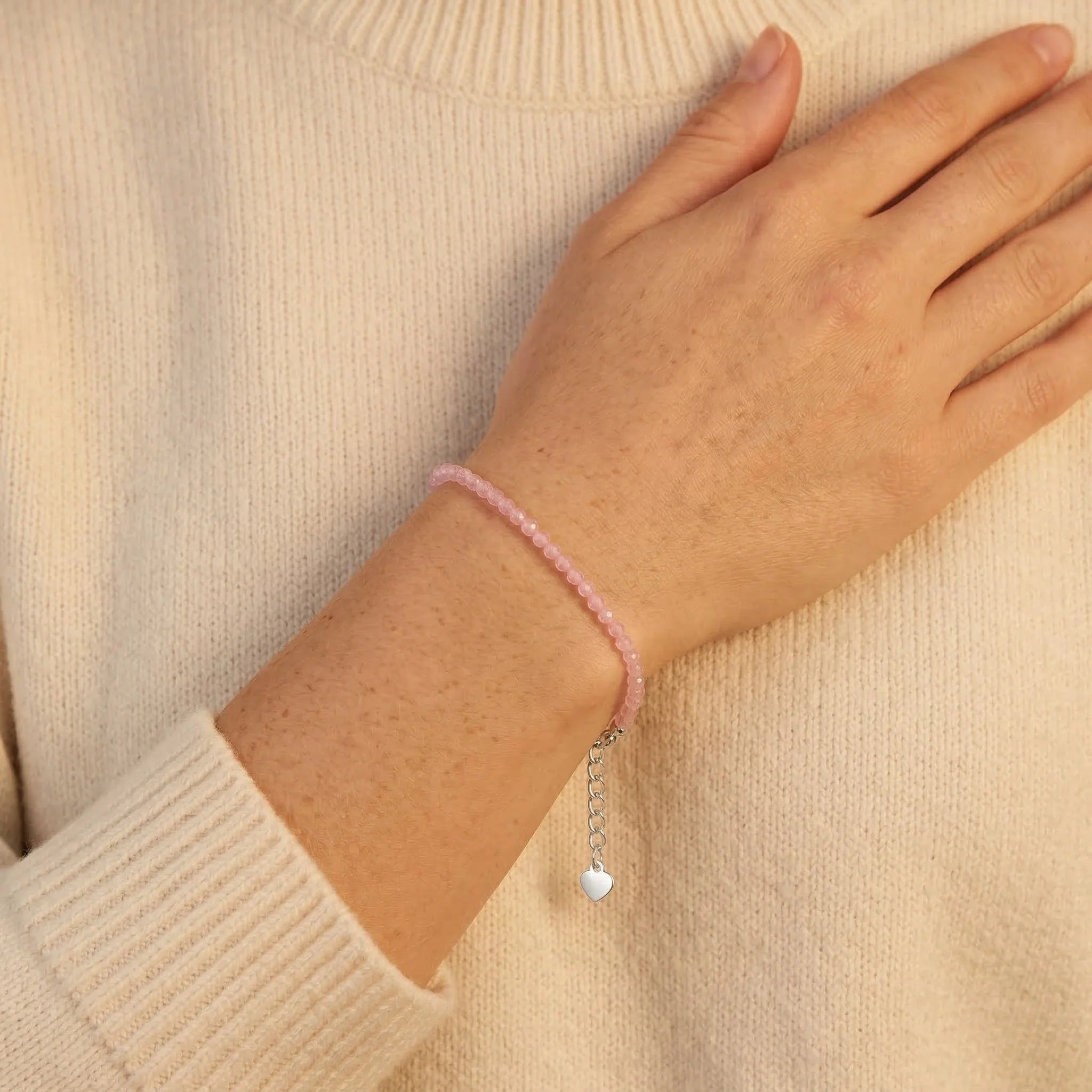 Hand wearing a pink beaded bracelet with a heart charm on a beige sweater.