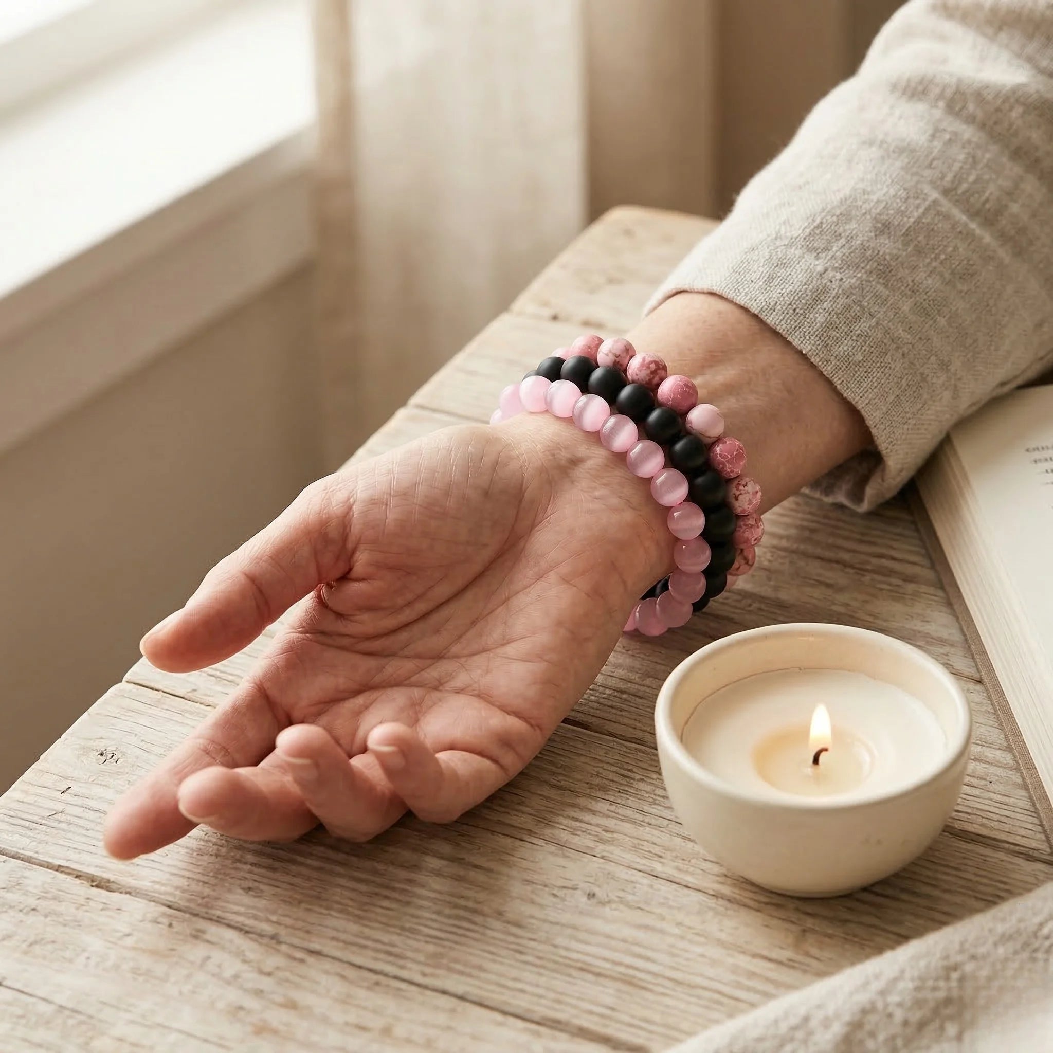 Hand with pink and black beaded bracelets resting on a wooden surface next to a small candle.