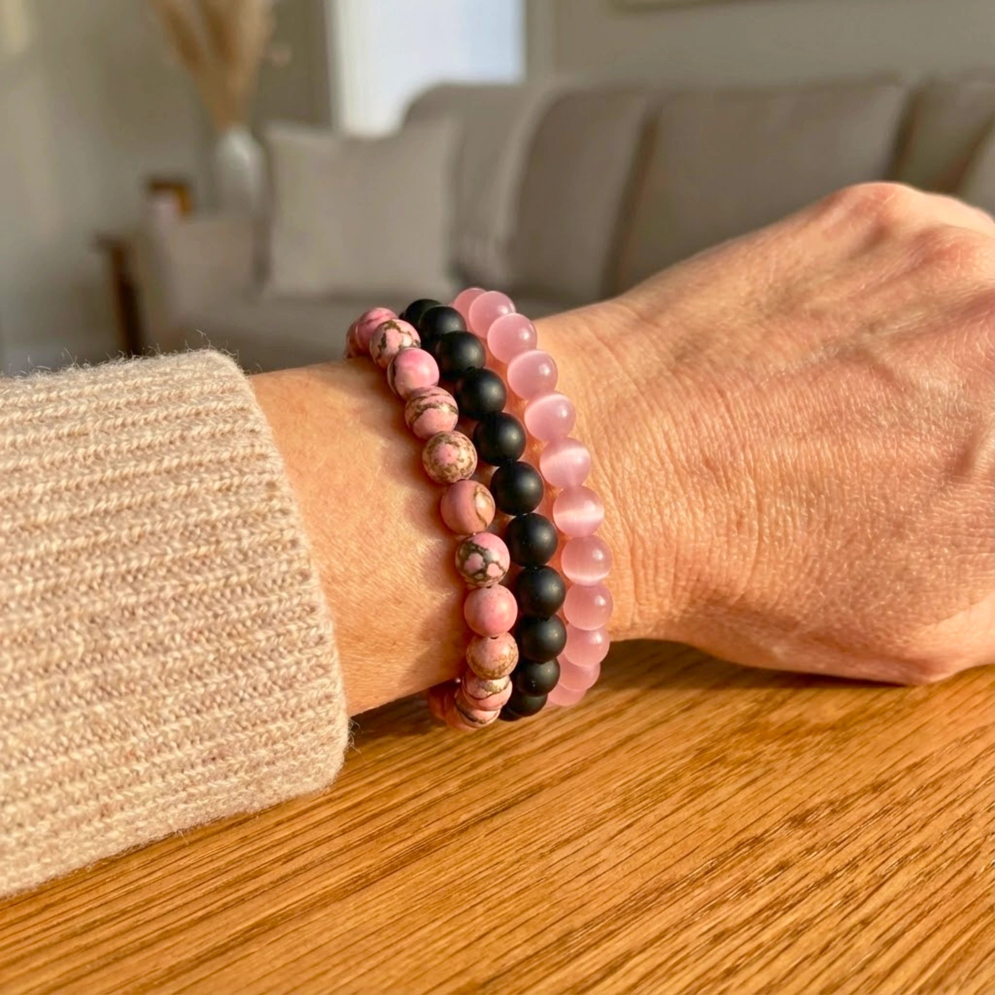 Person wearing two beaded Benevolence intention bracelet stack with rose pink quartz, black onyx, and pink imitation turquoise beads for love and kindness bracelets on a wooden surface with a blurred indoor background