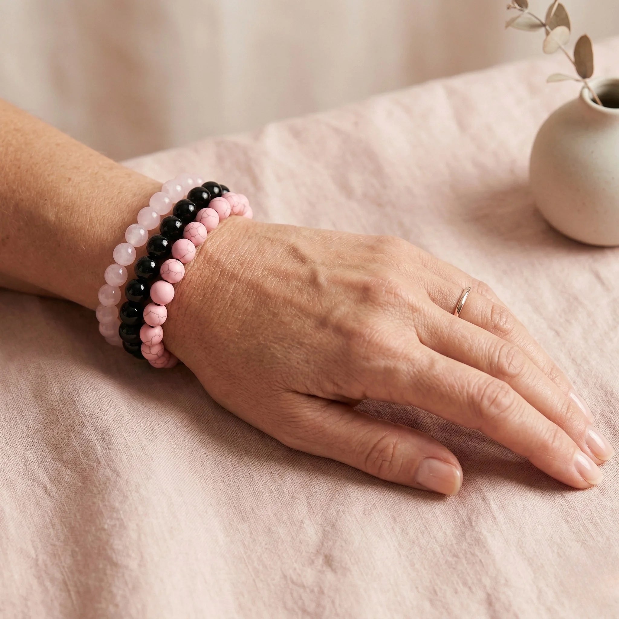 Hand wearing a pink and black beaded bracelet on a soft surface with a small vase in the background.