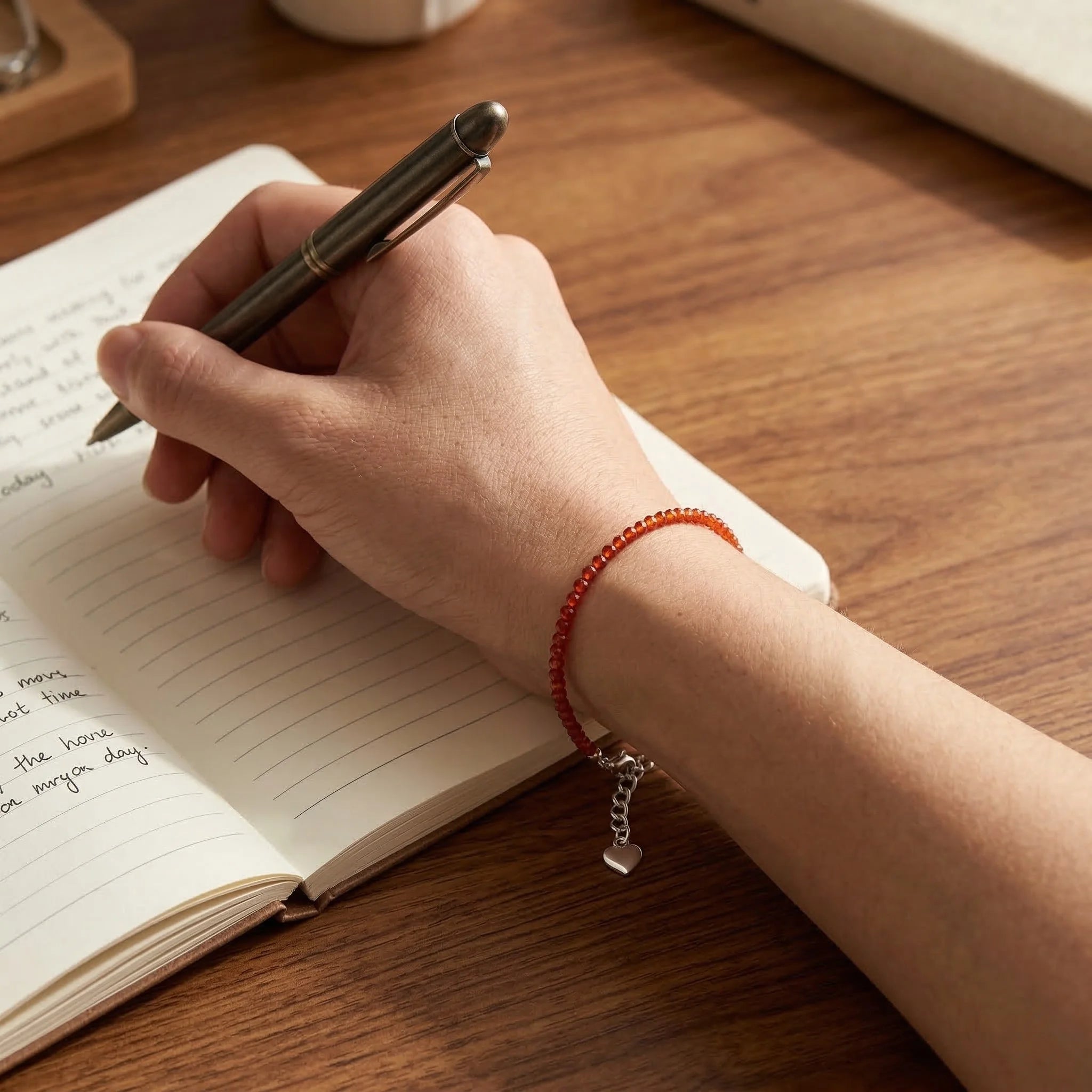 Hand writing in a notebook with a pen on a wooden surface, wearing a red beaded bracelet.