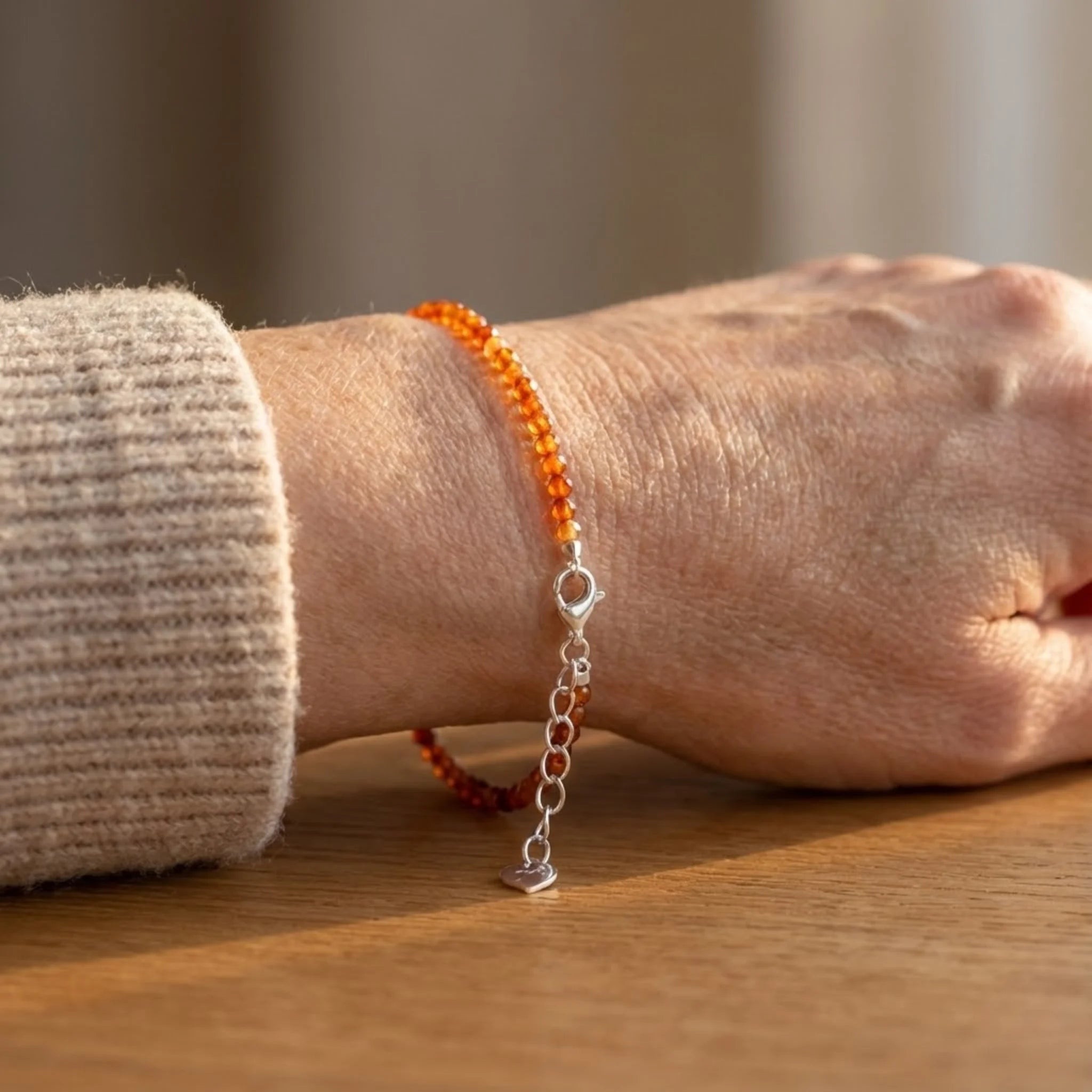Close-up of a wrist wearing an orange beaded Essential silver bracelet featuring garnet gemstone beads with rich red tones for subtle warmth bracelet with a silver chain on a wooden surface.