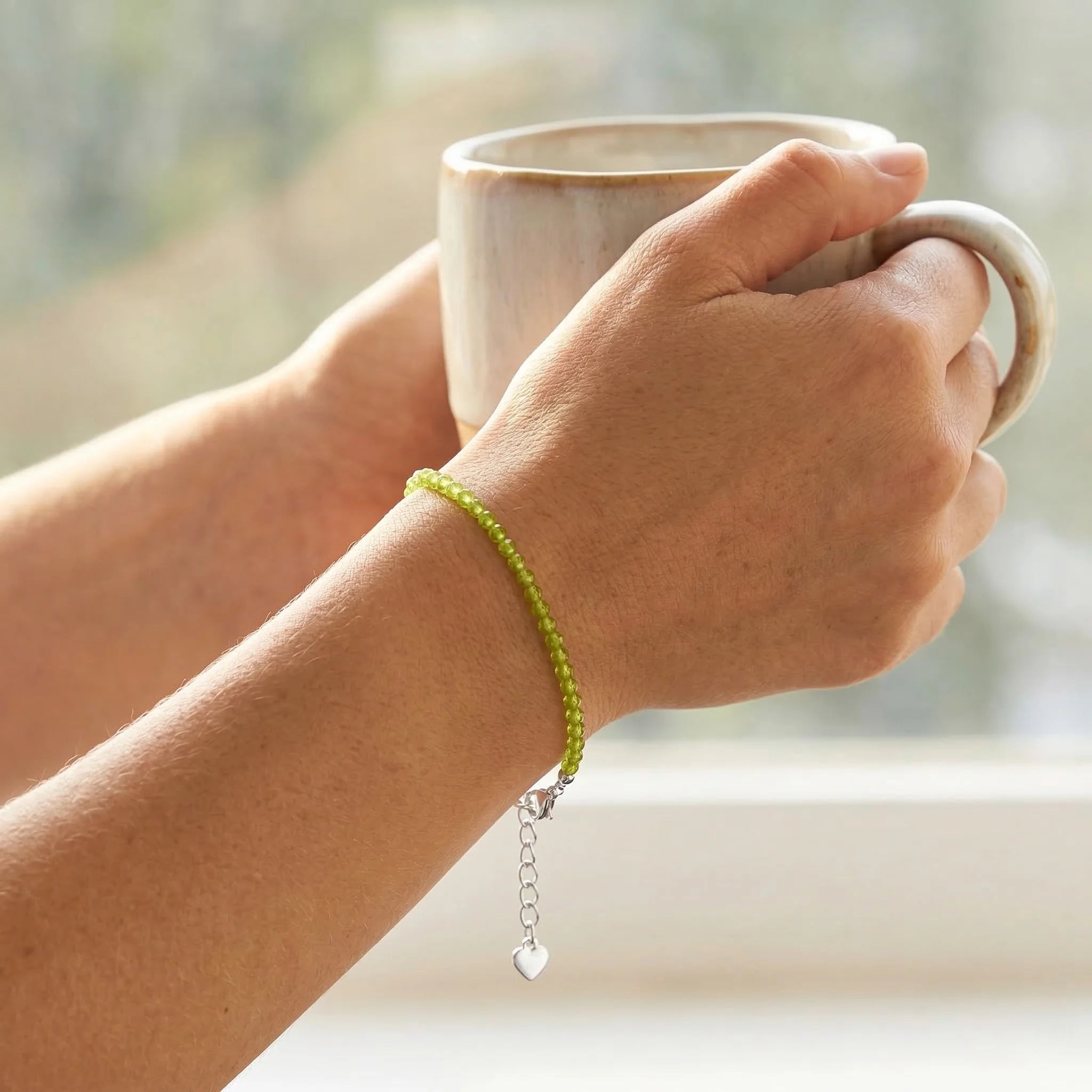 Hand holding a mug with a green beaded bracelet on a blurred natural background