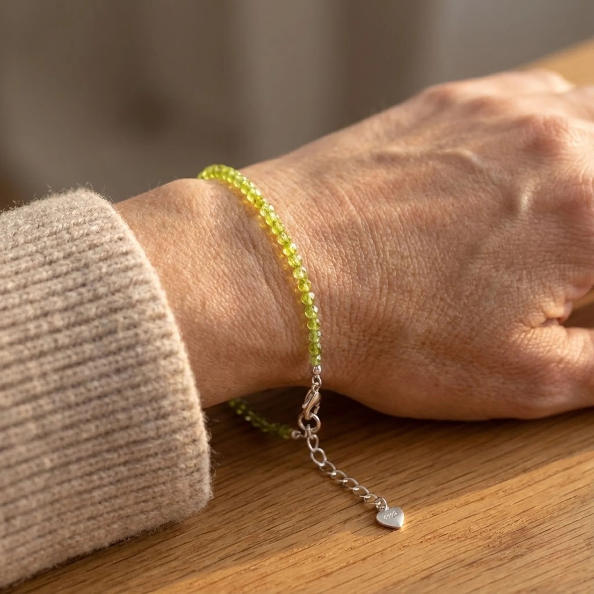 Person wearing a green beaded Essential silver bracelet made with peridot gemstone beads for a fresh, light green accent bracelet with a heart charm on a wooden surface