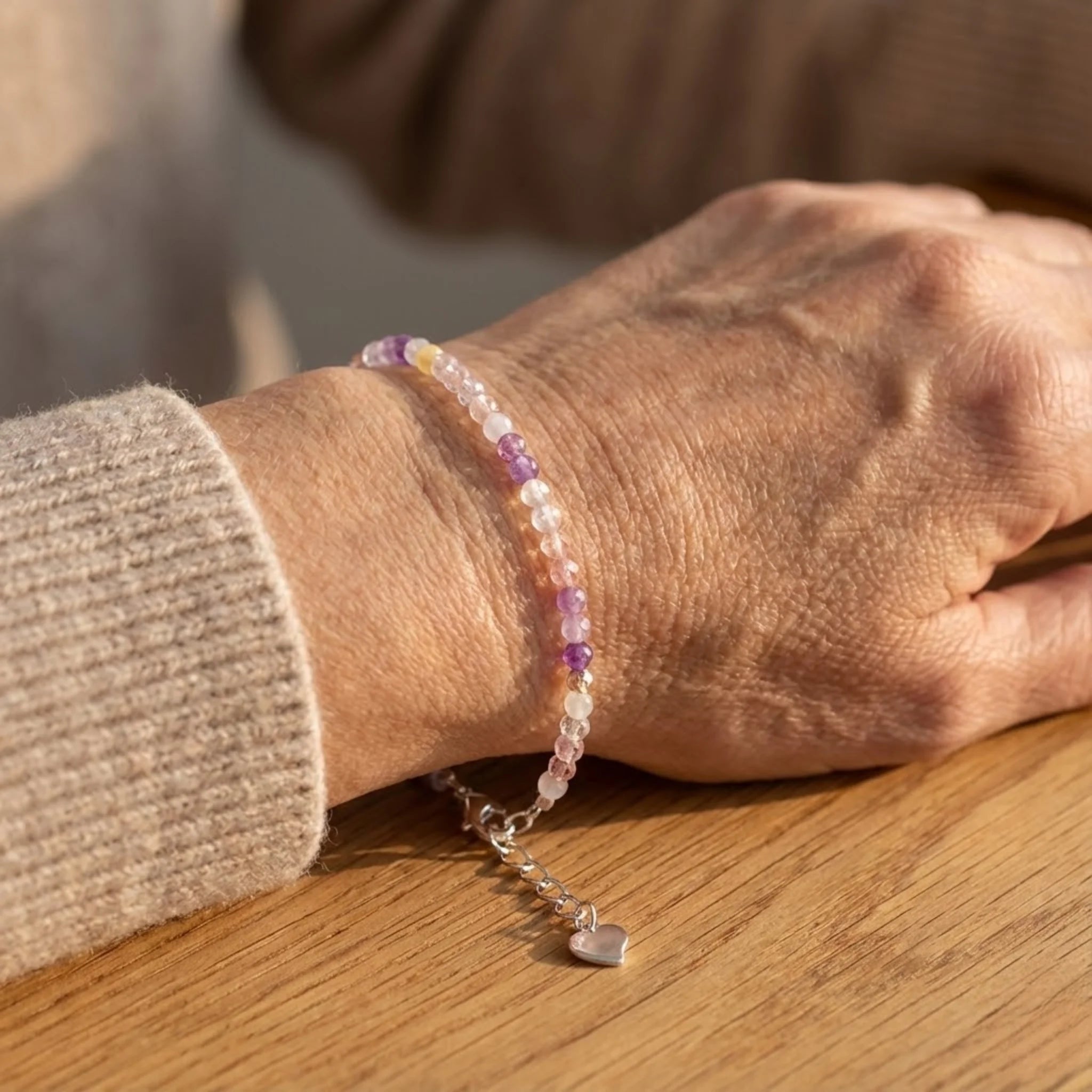 Close-up of a wrist wearing a beaded Essential silver bracelet made with purple quartz beads showing soft purple tones bracelet with a heart charm on a wooden surface.