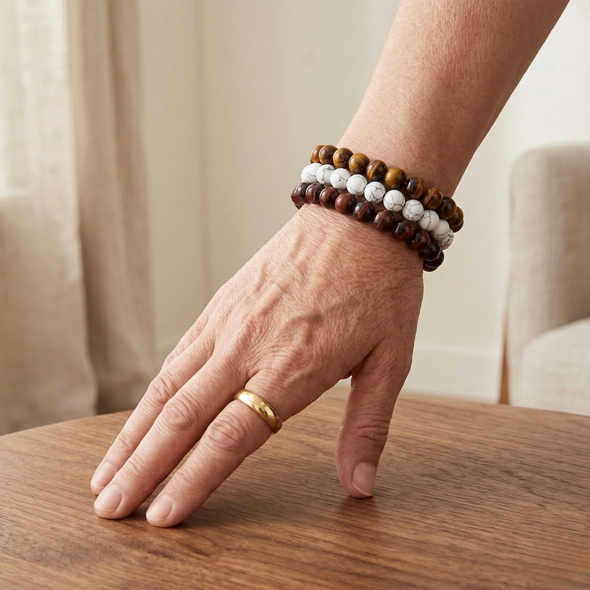 Hand wearing a gold ring and two beaded bracelets on a wooden surface with a neutral background