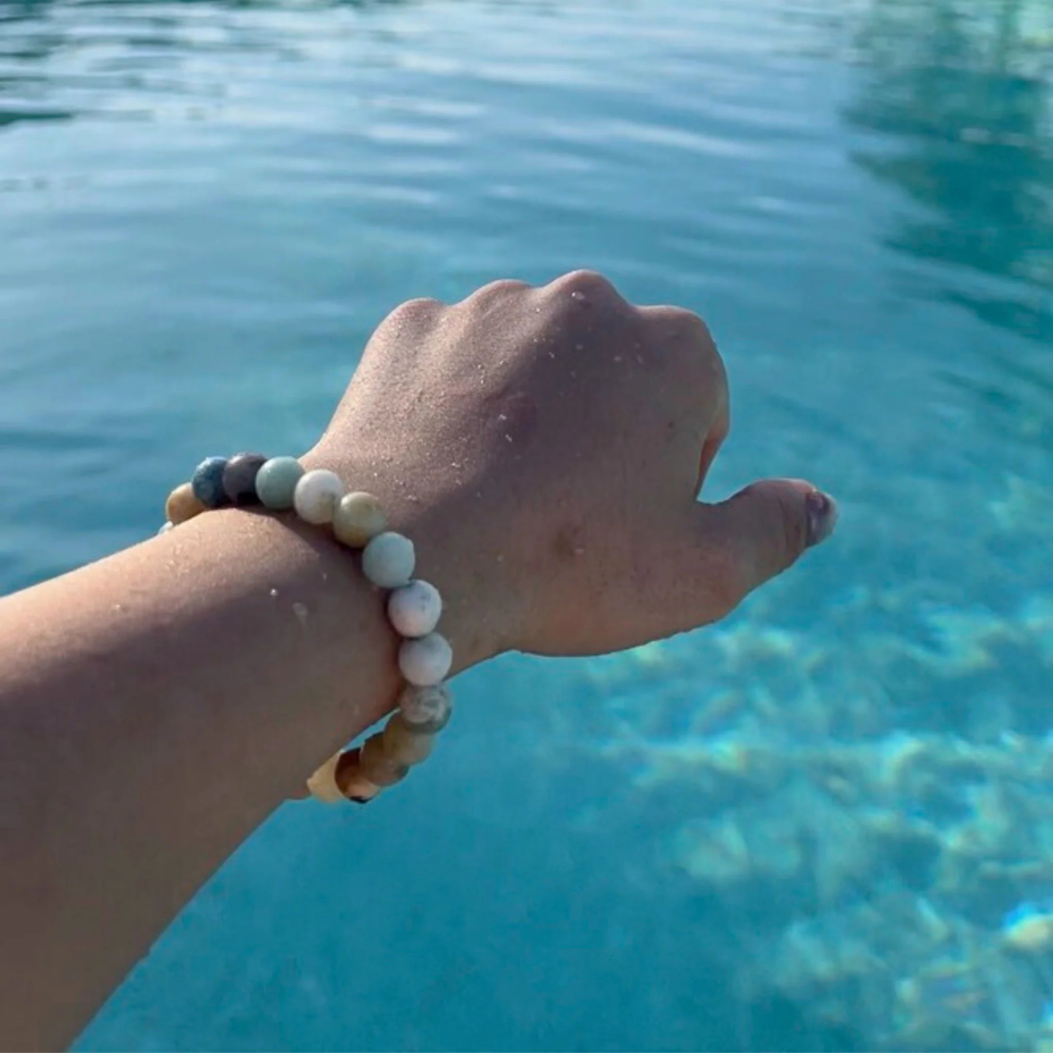 Gemstone4K amazon stone bracelet worn on wrist near beach or sunlight, showing aqua tones and reflective polish.